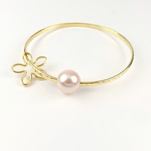 May include: A gold bangle bracelet with a delicate flower charm and a single pink pearl. The bracelet has a hammered texture.