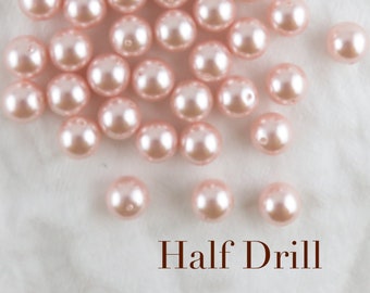 May include: A collection of pale pink, half-drilled faux pearls. The pearls are round with a glossy finish, scattered across a white surface. The text "Half Drill" is displayed in a dark brown font at the bottom of the image.