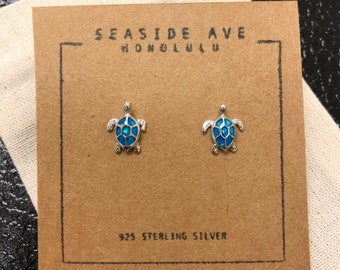 May include: A pair of sterling silver earrings featuring a blue turtle design. The earrings are displayed on a brown card with the text "SEASIDE AVE HONOLULU" and "925 STERLING SILVER".