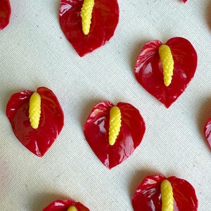 Puede incluir: Varios objetos rojos brillantes en forma de corazón con centros amarillos, que se asemejan a flores de anturio, están dispuestos sobre una superficie blanca. Los vibrantes pétalos rojos contrastan con el centro amarillo texturizado. Probablemente sean elementos decorativos o artesanales.