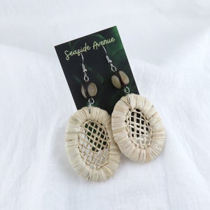 May include: A pair of woven, oval-shaped earrings with a natural, light brown color. The earrings are made of a woven material and have a small, round, dark brown bead at the top of each earring.