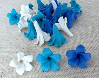 May include: A collection of handmade polymer clay flowers in shades of white, light blue, and dark blue. The flowers are in various stages of completion, with some fully formed and others still in bud form. The flowers are arranged on a neutral-colored surface.