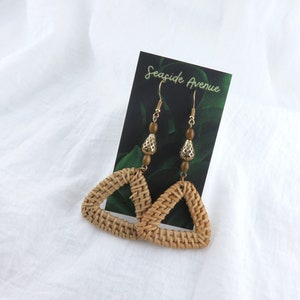 May include: A pair of woven triangle earrings with gold accents and brown beads. The earrings are hanging from a card that says "Seaside Avenue".