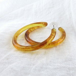 May include: A pair of large, amber-colored hoop earrings. The earrings are made of a translucent material with a marbled effect. The earrings are shown against a white background.