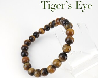 May include: A brown and gold tiger's eye beaded bracelet. The bracelet is made of round beads and is on a clear display stand. "Tiger's Eye" is written in green text above the bracelet.