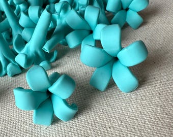May include: A collection of turquoise-colored, handmade clay flowers and succulent-like shapes. The flowers have rounded petals, and the succulents have elongated, curved forms. The objects are arranged on a textured, neutral-toned surface.