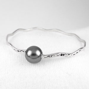 May include: A silver-toned bracelet with a wavy design and a dark gray pearl in the center. The bracelet has a hammered texture and is set against a white background. A piece of jewelry.
