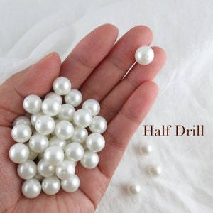 May include: A hand holding a pile of white pearl beads. The beads are round and smooth, and they have a slight shine. The text "Half Drill" is visible in the image.