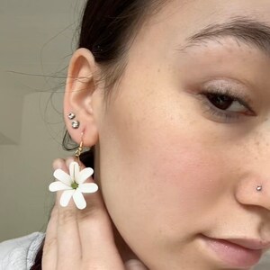 May include: A close-up of a person wearing a pair of white daisy flower earrings with gold-colored hooks. The earrings are held up by the person's hand. The person has a nose piercing and multiple ear piercings.