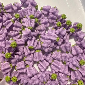 May include: A collection of small, lavender-colored flower-shaped beads with green accents. The beads are clustered together, showcasing their detailed floral design. These beads are likely used for jewelry making or other craft projects.
