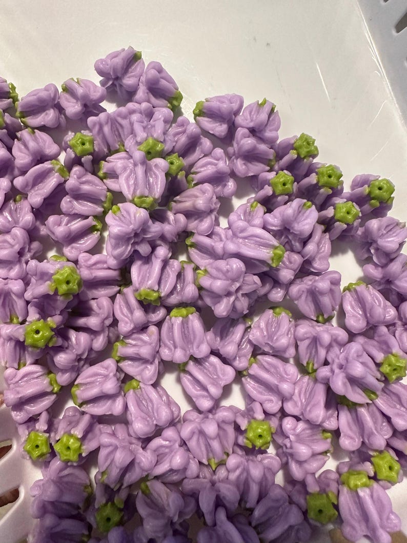 May include: A collection of small, lavender-colored flower-shaped beads with green accents. The beads are clustered together, showcasing their detailed floral design. These beads are likely used for jewelry making or other craft projects.