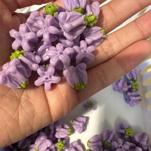 May include: A collection of small, light purple flower-shaped beads with green centers. Each bead features five petals arranged in a bell-like structure, with a small hole in the center for stringing. The beads have a smooth, matte finish.