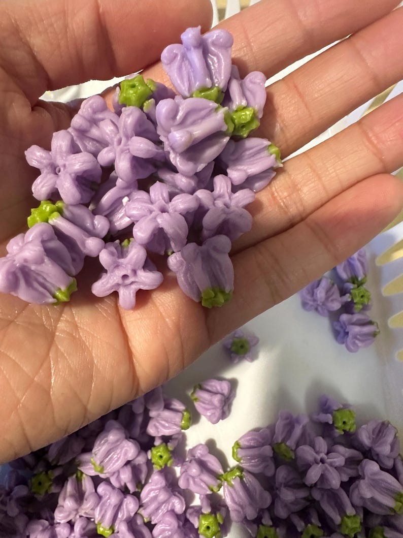 May include: A collection of small, light purple flower-shaped beads with green centers. Each bead features five petals arranged in a bell-like structure, with a small hole in the center for stringing. The beads have a smooth, matte finish.