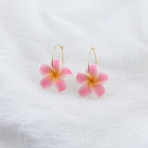 Puede incluir: Un par de pendientes de aro dorados con dijes de flores rosas. Las flores tienen un centro blanco y p&eacute;talos rosas.