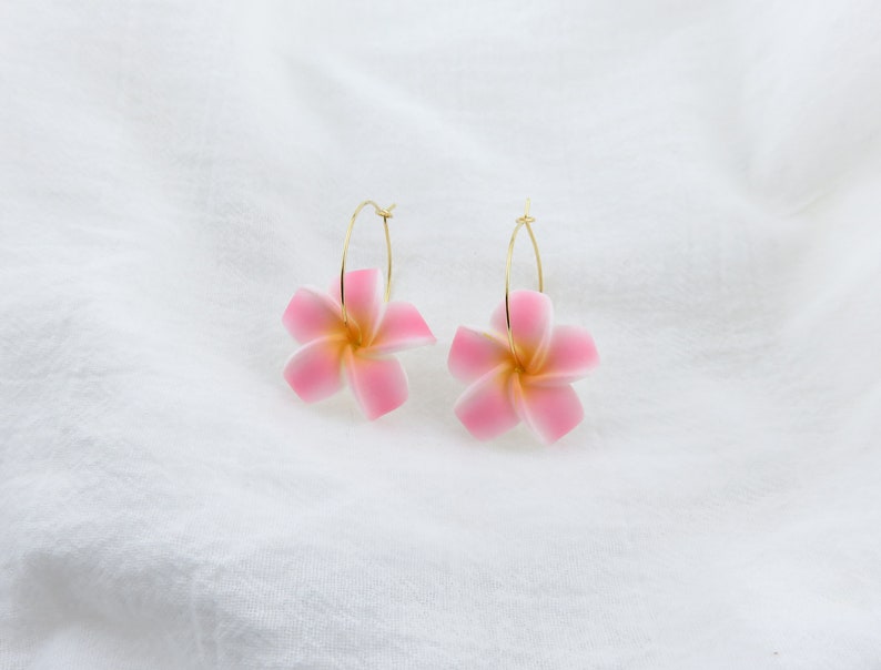 Puede incluir: Un par de pendientes de aro dorados con dijes de flores rosas. Las flores tienen un centro blanco y p&eacute;talos rosas.