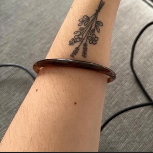 May include: A smooth, translucent dark brown bangle bracelet with a rounded profile. The bracelet is worn on a forearm that features a black ink tattoo of a small bouquet of wildflowers tied together with a simple string at the stems.