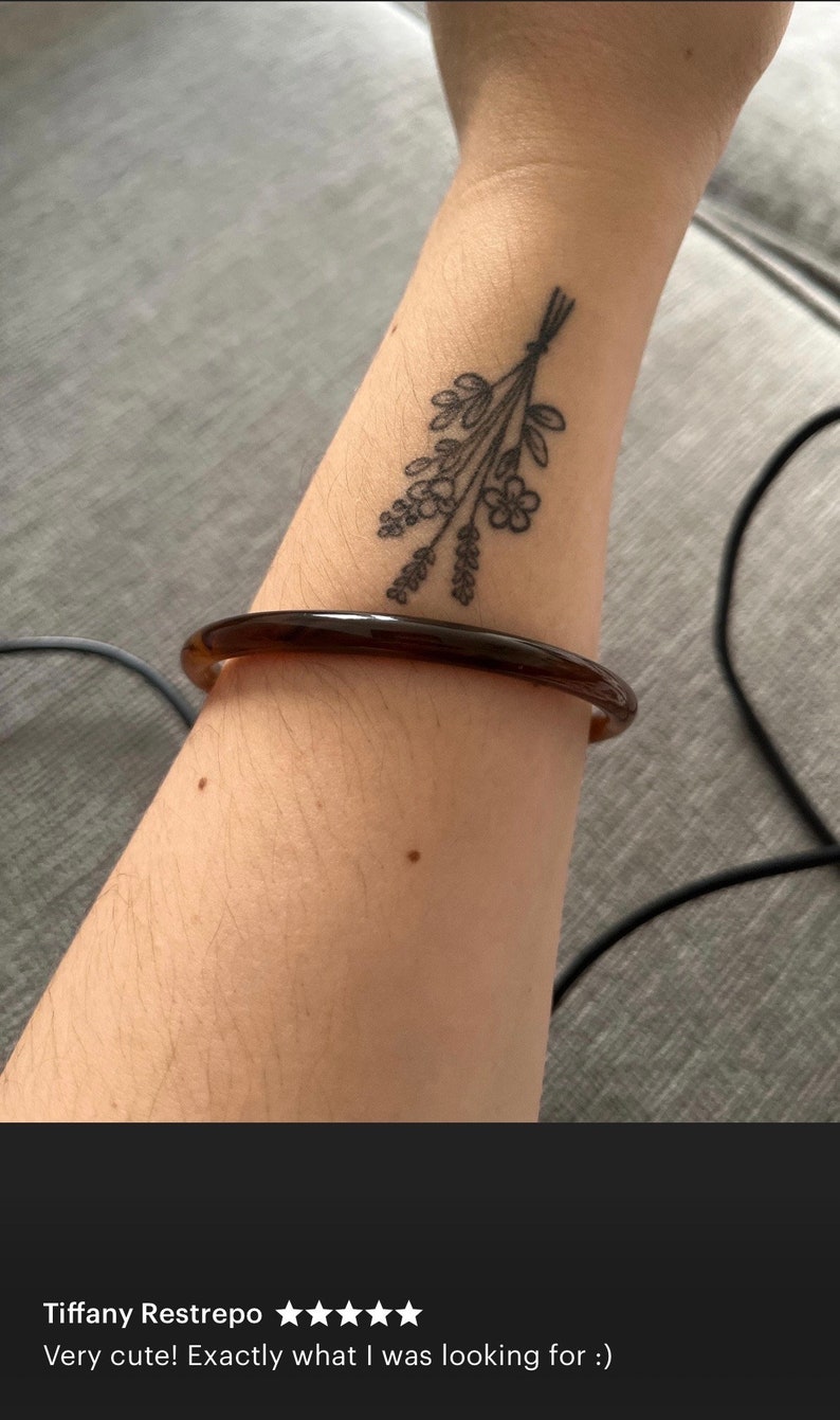 May include: A smooth, translucent dark brown bangle bracelet with a rounded profile. The bracelet is worn on a forearm that features a black ink tattoo of a small bouquet of wildflowers tied together with a simple string at the stems.