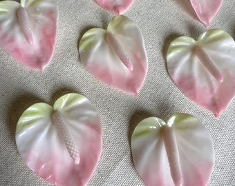 May include: A collection of ceramic anthurium flower-shaped decorations. Each piece features a gradient of colors, transitioning from a pale green at the base to a soft pink at the tips. The center of each flower has a textured, light pink spadix.