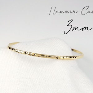 May include: A gold hammered bangle bracelet with a 3mm width. The bracelet is on a white fabric background.