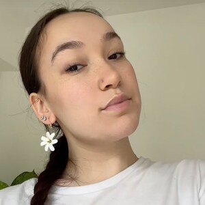 May include: White flower earrings with green accents. The earrings are in the shape of a flower with multiple petals. The earrings are worn by a person with dark hair and a white t-shirt.