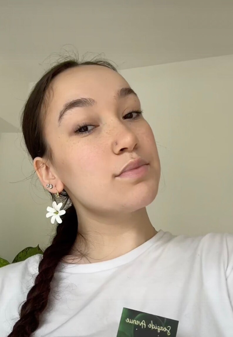 May include: White flower earrings with green accents. The earrings are in the shape of a flower with multiple petals. The earrings are worn by a person with dark hair and a white t-shirt.