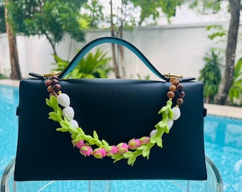 May include: A black handbag with a teal handle and a decorative floral strap. The strap features green leaves, white flower buds, and pink rosebuds. The bag is sitting in front of a pool.