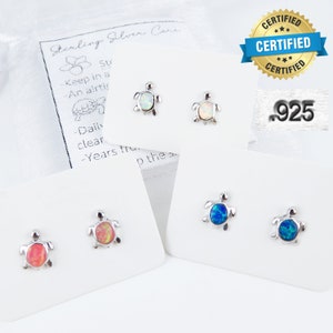 May include: Four pairs of sterling silver turtle stud earrings with opal stones. The earrings are displayed on white cards. The stones are pink, white, blue, and light blue. The cards have the text "Sterling Silver Care" and "925" printed on them. The earrings are certified.