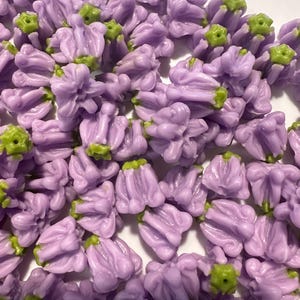May include: A collection of small, lavender-colored flower-shaped beads with green accents. These beads are clustered together, showcasing their detailed floral design. Ideal for jewelry making or craft projects.
