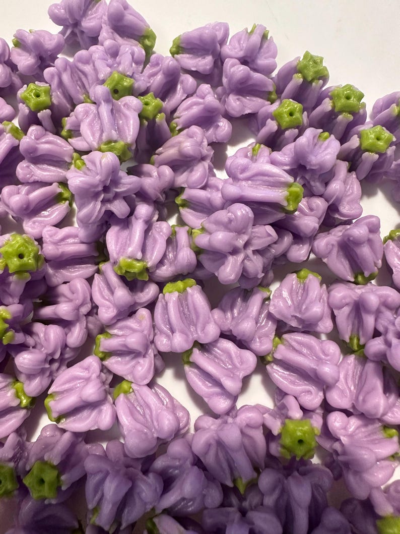 May include: A collection of small, lavender-colored flower-shaped beads with green accents. These beads are clustered together, showcasing their detailed floral design. Ideal for jewelry making or craft projects.