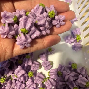 May include: A collection of small, lavender-colored flower beads with green accents. The beads are clustered together, some held in a hand, and others scattered in a white basket. These decorative beads are ideal for crafting and jewelry making.