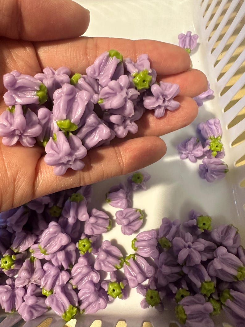 May include: A collection of small, lavender-colored flower beads with green accents. The beads are clustered together, some held in a hand, and others scattered in a white basket. These decorative beads are ideal for crafting and jewelry making.