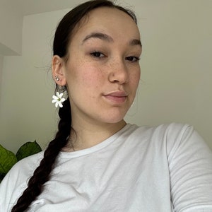 May include: White flower earrings with a green center are worn on the left ear. The earrings are hanging from a gold hoop. The person is wearing a white t-shirt and has dark brown hair in a braid.