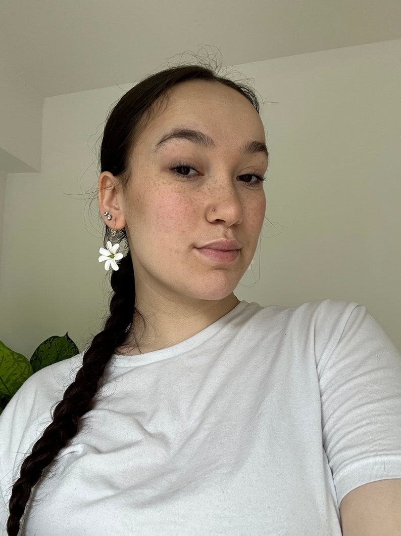 May include: White flower earrings with a green center are worn on the left ear. The earrings are hanging from a gold hoop. The person is wearing a white t-shirt and has dark brown hair in a braid.