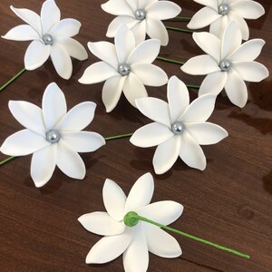 May include: Eight white paper flowers with silver centers and green stems. The flowers are arranged on a brown wooden surface.