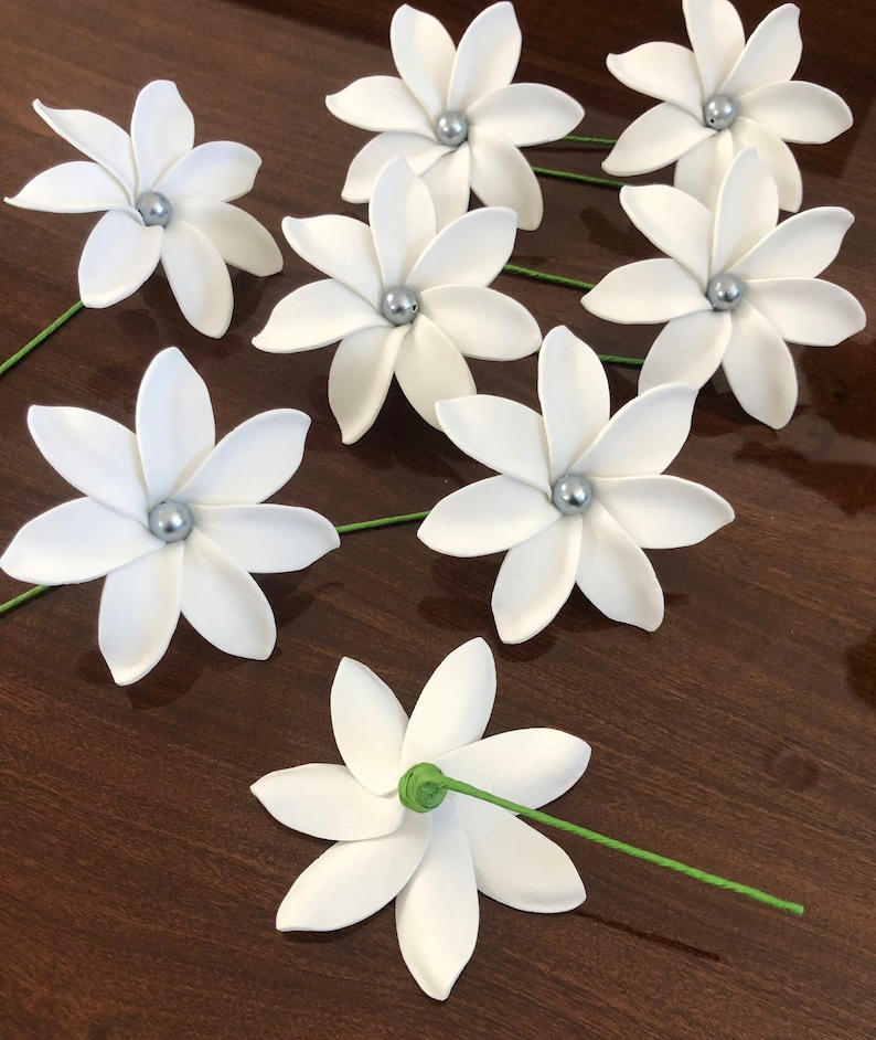 May include: Eight white paper flowers with silver centers and green stems. The flowers are arranged on a brown wooden surface.