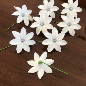 May include: Eight white paper flowers with silver centers and green stems. The flowers are arranged on a brown wooden surface.