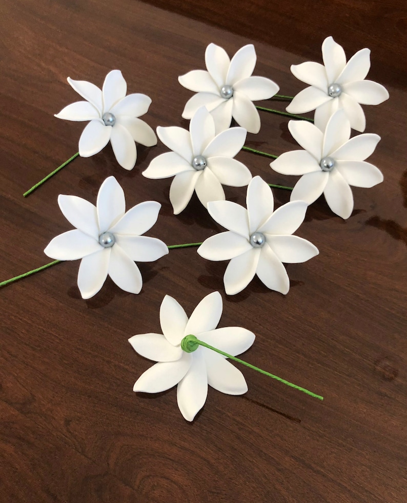 May include: Eight white paper flowers with silver centers and green stems. The flowers are arranged on a brown wooden surface.