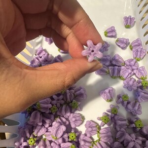 May include: A collection of small, lavender-colored flower beads with green accents. The beads are held in a white plastic basket. One bead is held up by a hand, showcasing its star-like shape and central hole. These beads are ideal for jewelry making.