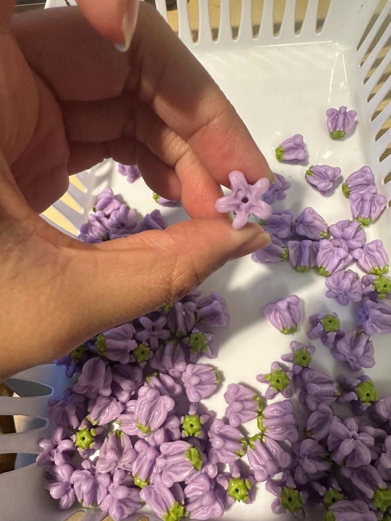 May include: A collection of small, lavender-colored flower beads with green accents. The beads are held in a white plastic basket. One bead is held up by a hand, showcasing its star-like shape and central hole. These beads are ideal for jewelry making.