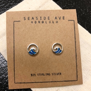 Puede incluir: Un par de pendientes de plata de ley con un diseño de ola azul. Los pendientes están empaquetados en una tarjeta marrón con el texto "Seaside Ave Honolulu" y "925 Sterling Silver".