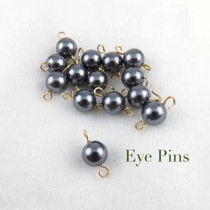 May include: A group of 15 dark gray eye pins with gold loops. The eye pins are scattered on a white surface. The text "Eye Pins" is written in green below the eye pins.
