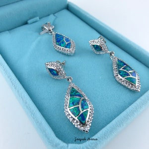 May include: A set of silver earrings and a pendant featuring a teardrop-shaped design with blue and green opal stones and clear cubic zirconia accents. The jewellery is displayed in a light blue box.