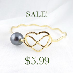 May include: Gold bangle bracelet with a heart-shaped infinity symbol and a black pearl. The bracelet is on a white background. SALE! $5.99