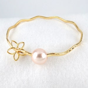 May include: A gold bangle bracelet with a delicate flower charm and a single pink pearl. The bracelet has a hammered texture.