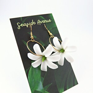 May include: A pair of gold-tone hoop earrings with white flower pendants. The earrings are displayed on a black card with the text "Seaside Avenue" and a green leaf design. The earrings have a delicate and feminine design.
