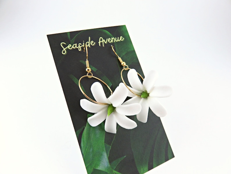 May include: A pair of gold-tone hoop earrings with white flower pendants. The earrings are displayed on a black card with the text "Seaside Avenue" and a green leaf design. The earrings have a delicate and feminine design.