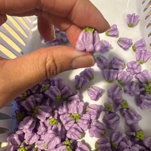 May include: A hand holding a small, lavender-colored flower bead with a green base. The bead is one of many similar beads in a white basket. The beads are likely for jewelry making or other crafts.