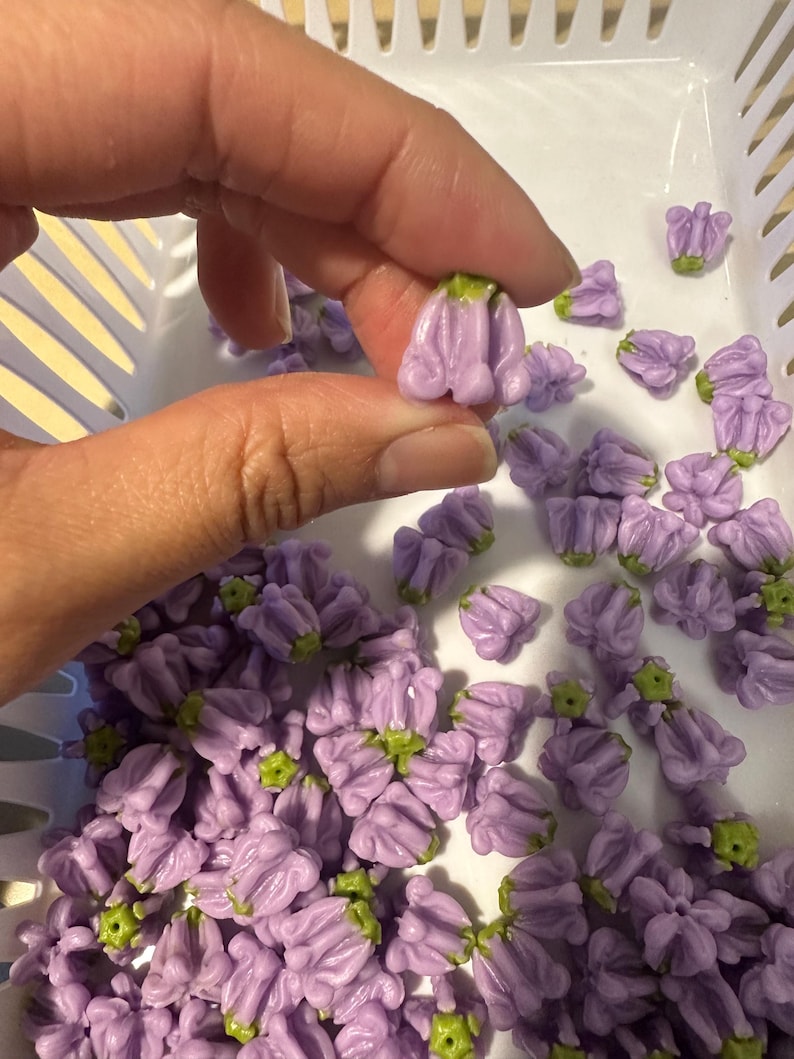 May include: A hand holding a small, lavender-colored flower bead with a green base. The bead is one of many similar beads in a white basket. The beads are likely for jewelry making or other crafts.