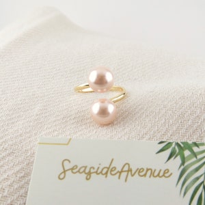 以下が含まれることがあります： 2つのピンクの真珠のアクセントが付いたゴールドリング。リングはねじれており、繊細なデザインが施されています。背景のカードにはSeaside Avenueと印刷されています。