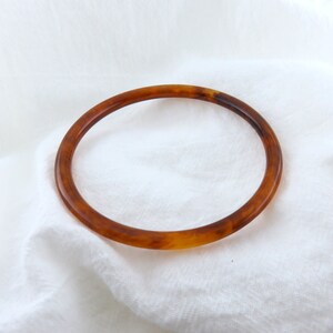 May include: A circular bangle bracelet featuring a translucent amber and brown tortoiseshell pattern. The bracelet has a smooth, polished finish and a rounded profile, showcasing a classic mottled design with varying shades of golden brown.